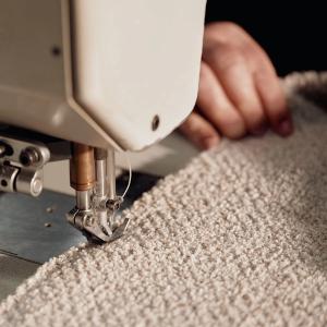 Dit is een close-up van een naaimachine die een zachte beige stof naait, wat het ambachtelijke proces van het vervaardigen van meubels of stoffering benadrukt.