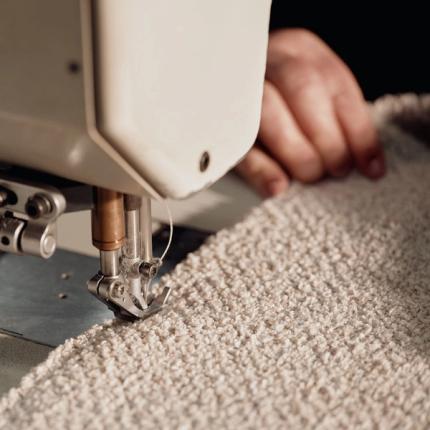 Dit is een close-up van een naaimachine die een zachte beige stof naait, wat het ambachtelijke proces van het vervaardigen van meubels of stoffering benadrukt.
