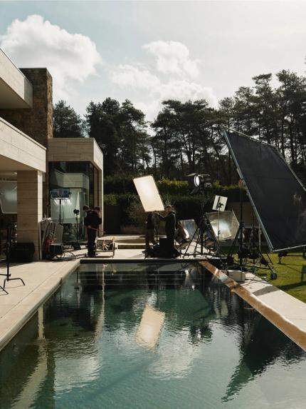 Coulisses d’un tournage près d’une maison moderne avec piscine ; l’équipe et de grands panneaux de diffusion sont installés dans le jardin.