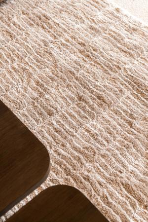 Detail van een beige hoogpolig tapijt met organisch reliëf naast de afgeronde randen van houten salontafels.