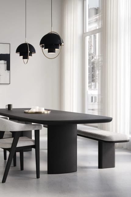 Bootvormige Amato eettafel in zwart hout met bijpassende bank en stoel in lichte stof, onder twee zwarte hanglampen bij een groot raam met witte gordijnen.