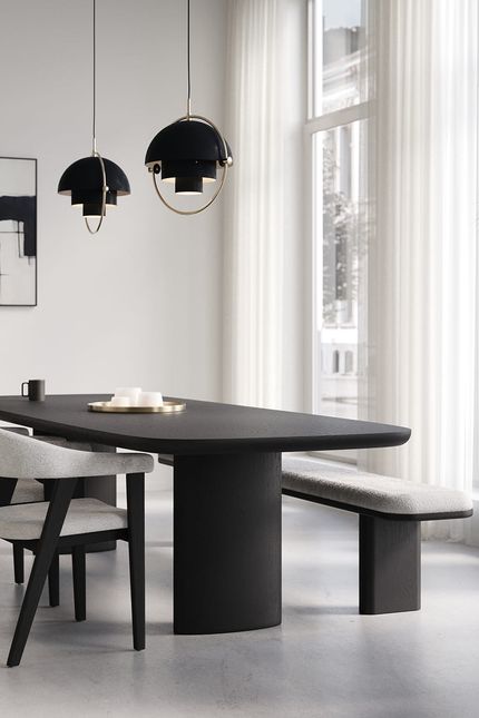 Bootvormige Amato eettafel in zwart hout met bijpassende bank en stoel in lichte stof, onder twee zwarte hanglampen bij een groot raam met witte gordijnen.