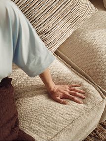Gros plan d’une main appuyant sur le revêtement doux et clair d’un canapé, avec un coussin décoratif rayé à l’arrière-plan.