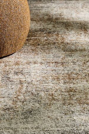 Detail van een gemêleerd tapijt in natuurlijke beige- en bruintinten naast een gestoffeerd meubel.