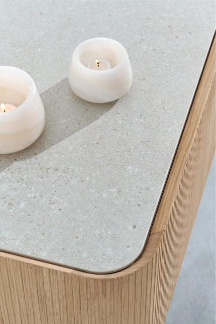 Close-up van een houten kast met verticale latjes en een licht, steenlook bovenblad met afgeronde hoeken, met twee witte kaarsenhouders.