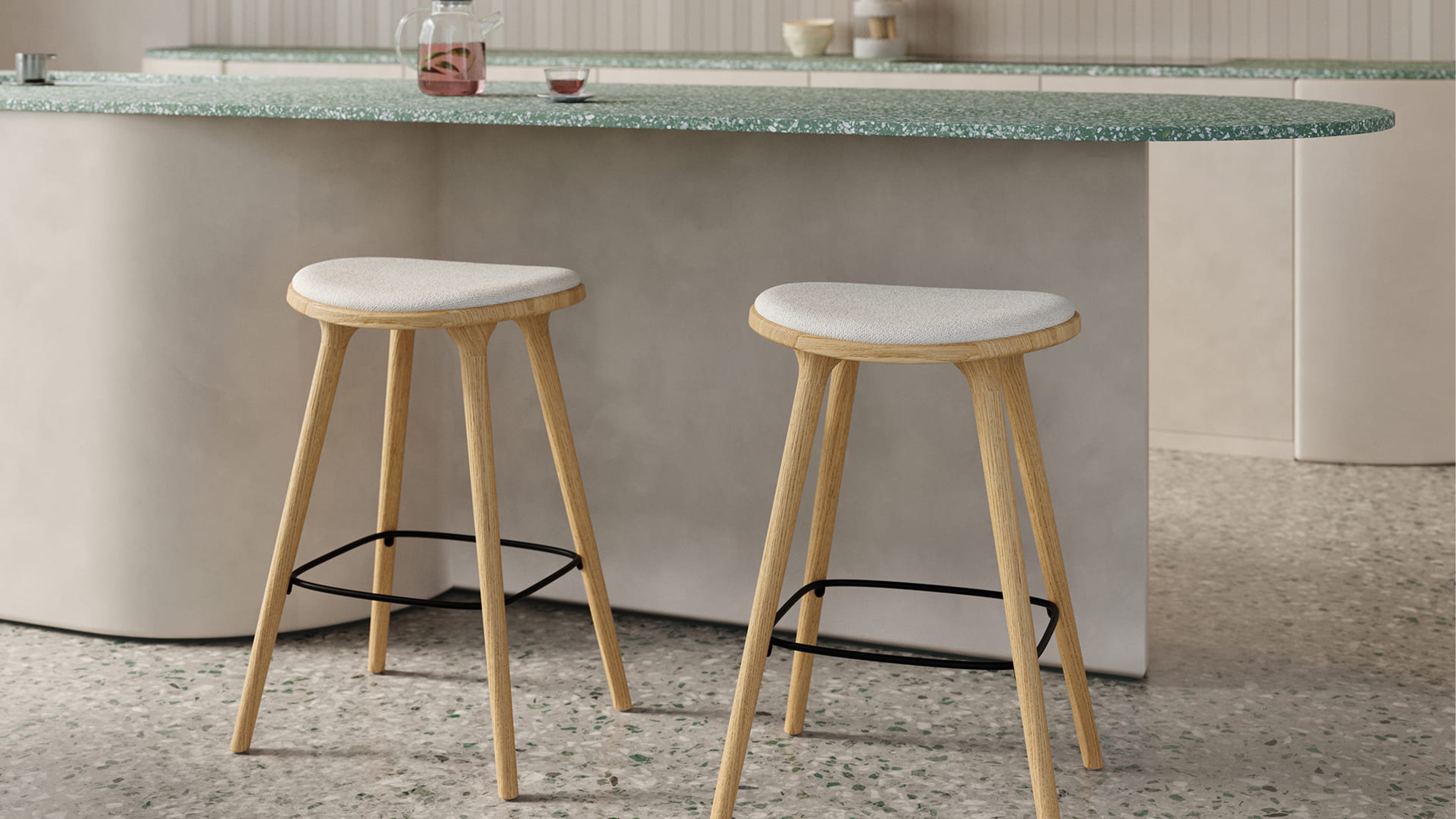 Twee houten barkrukken met licht gestoffeerde zitting onder een afgerond keukeneiland met groen gespikkeld blad, op een terrazzo vloer.