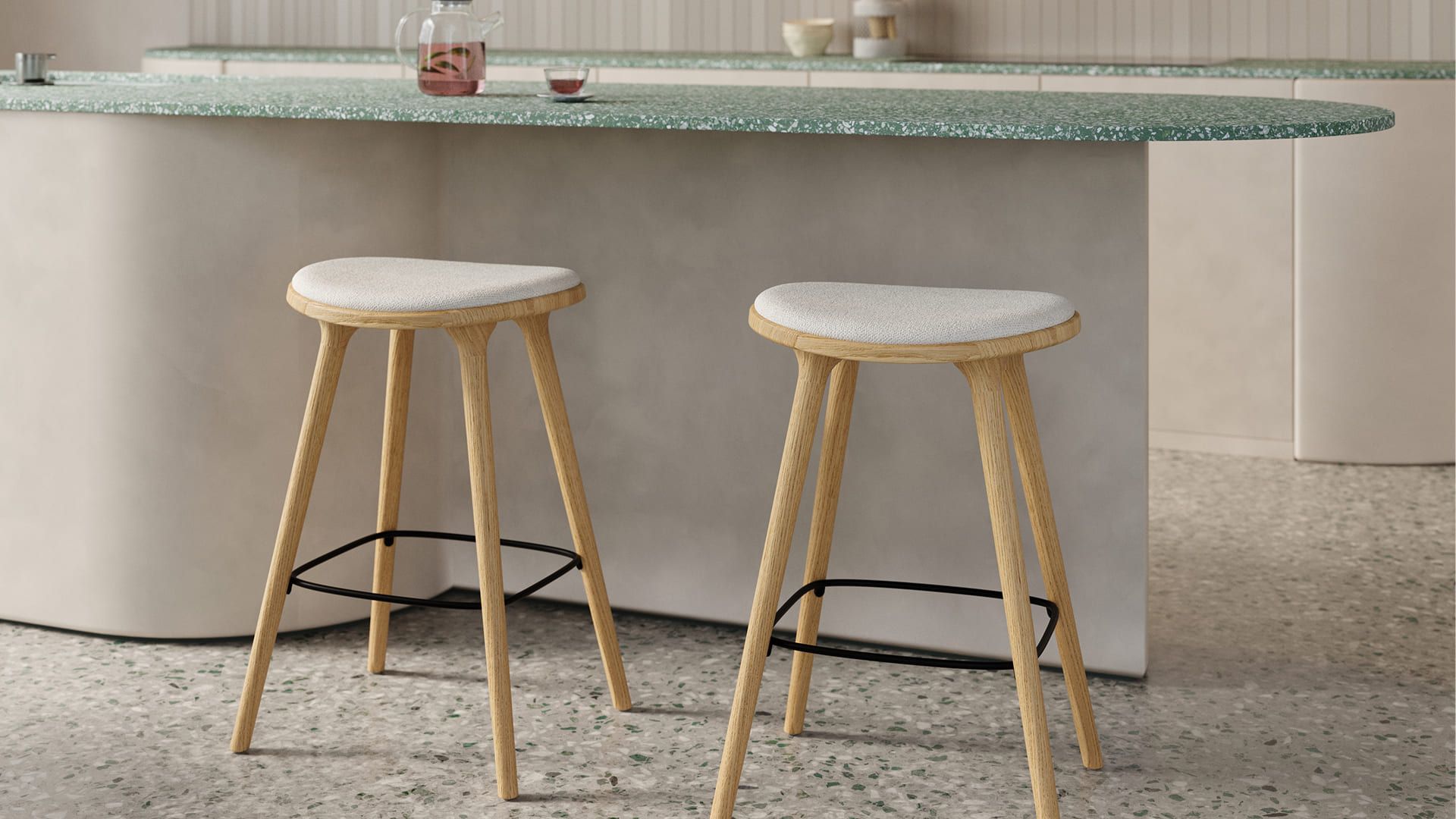 Twee houten barkrukken met licht gestoffeerde zitting onder een afgerond keukeneiland met groen gespikkeld blad, op een terrazzo vloer.