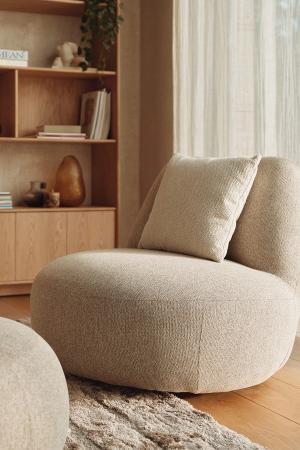 Comfortabele, ronde fauteuil met een beige stoffering en een kussen, voor een houten kast en een lichte, moderne ruimte.