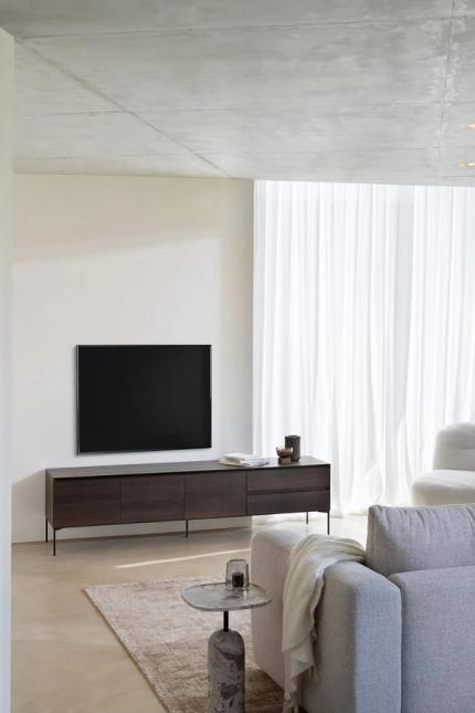 Meuble TV en bois sombre sur pieds fins sous une télévision murale, dans un salon lumineux avec des rideaux transparents.