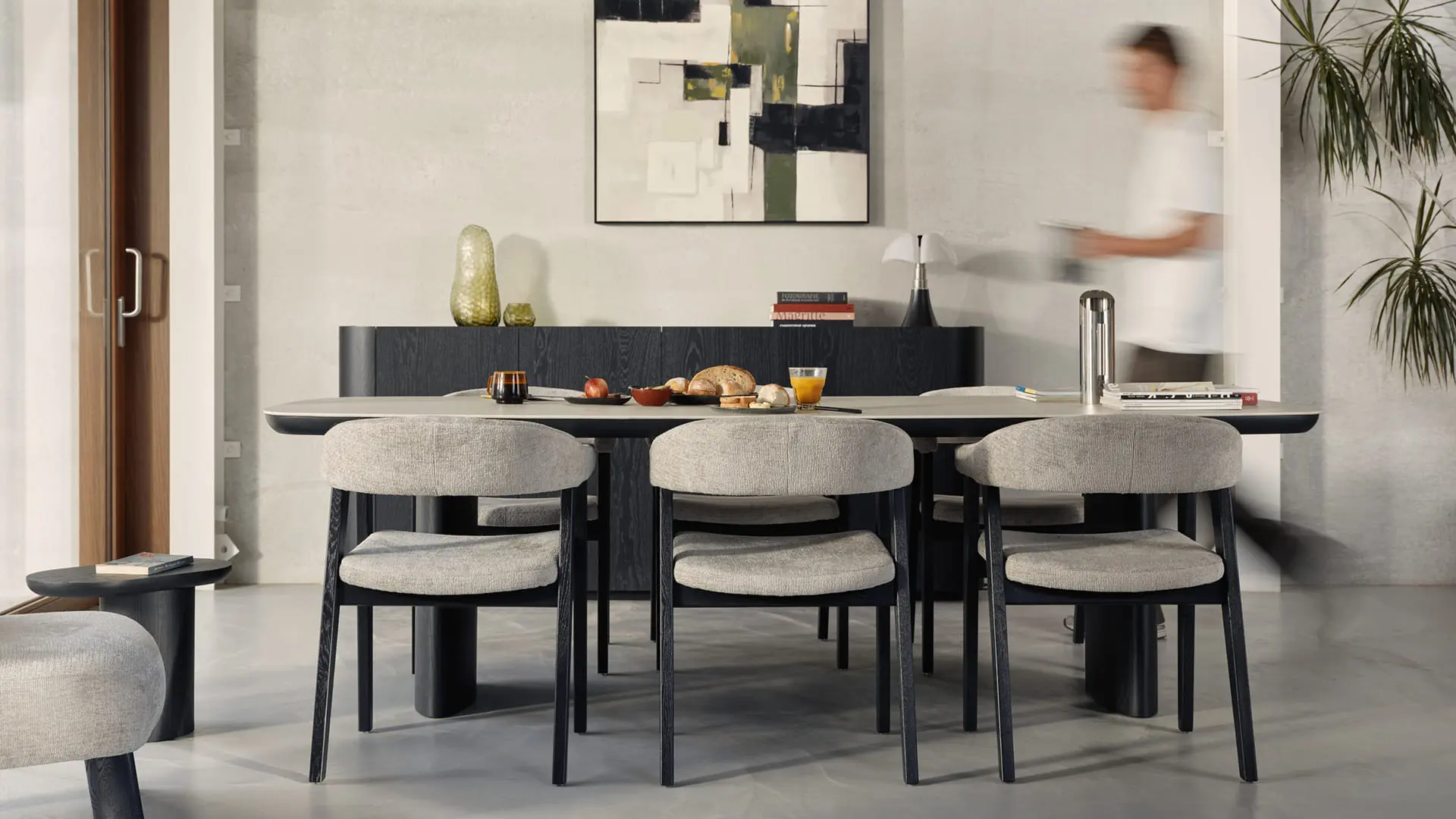 Lange eettafel met vier grijze stoelen en donkere poten, stilleven op tafel, abstract kunstwerk aan de muur en persoon in beweging.