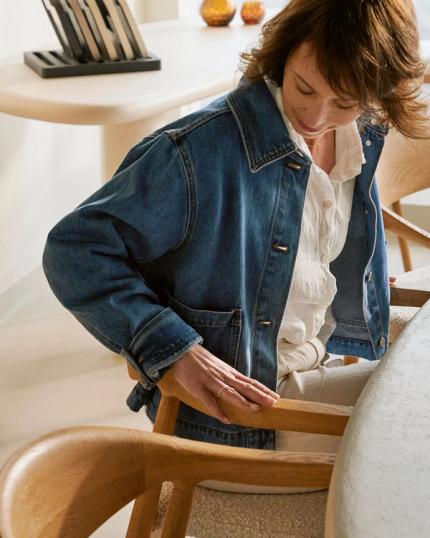 Une femme en veste en jean ajustant le dossier d'une chaise de salle à manger en bois dans un espace moderne et minimaliste.