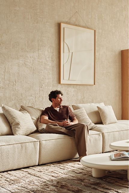 Man zit ontspannen op een lichte zetel in een rustige woonkamer met witte salontafels, groot raam en zacht daglicht.
