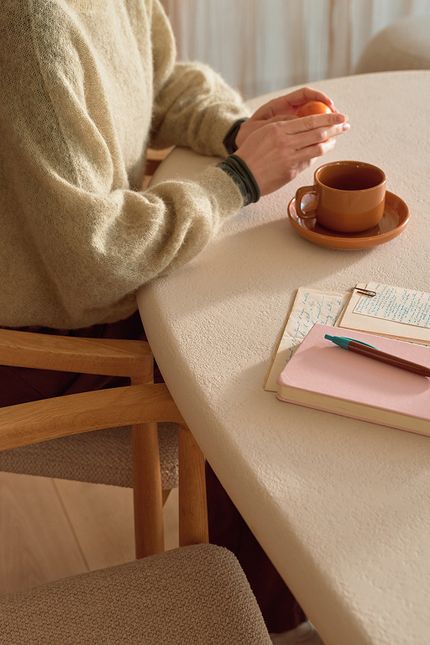 Een persoon zit aan een lichte, ronde eettafel, met een kopje koffie en een sinaasappel in de handen. Op de tafel liggen een roze notitieboek, een pen, en een stapel papieren met handgeschreven aantekeningen.
