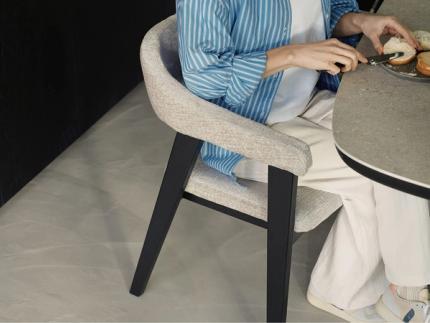 Une personne tartine du pain à table, assise sur une chaise rembourrée avec des pieds en bois noir.