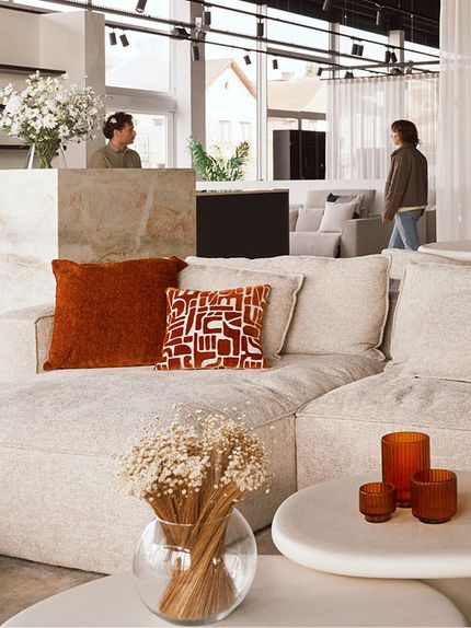 Showroom met een grote beige bank, oranjerode kussens en decoratieve objecten, een vaas met gedroogde bloemen op de salontafel, en een persoon die door de ruimte beweegt achter een marmeren balie.