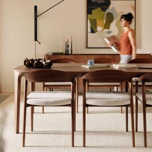 Curvo eettafel met houten stoelen, gestyled met een kunstzinnige schaal en boeken. Een vrouw loopt voorbij, met een schilderij en lamp op de achtergrond.