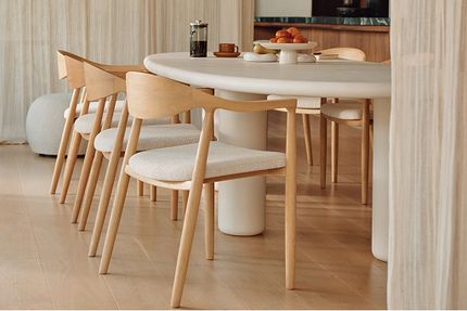 Close-up van een ovale eettafel met afgeronde rand en witte kolompoot, met lichte houten stoelen en gestoffeerde zittingen eromheen.