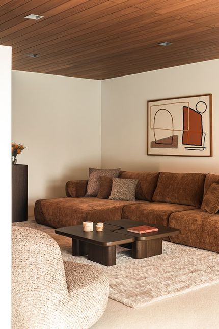 Warme woonkamer met een bruine hoekzetel, donkere salontafel en fauteuil in neutrale tinten.