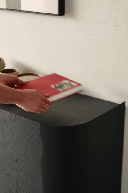 Gros plan d'une main posant un livre rouge sur un buffet en bois foncé avec un coin arrondi, contre un mur clair.