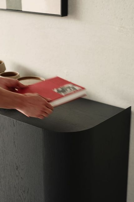 Gros plan d'une main posant un livre rouge sur un buffet en bois foncé avec un coin arrondi, contre un mur clair.